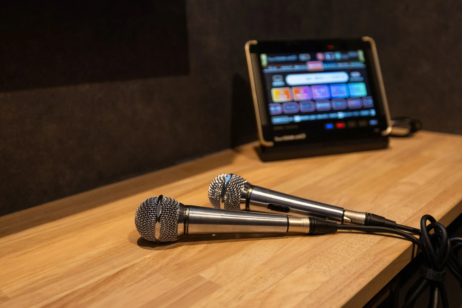 木目調のテーブルに2本のマイクが置かれ、奥にはカラオケの選曲画面らしきタブレットが見える