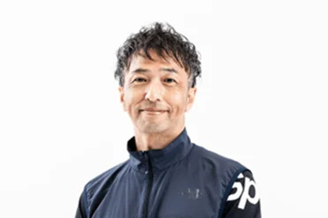 白い背景の前で、ダークカラーのスポーツウェアを着て微笑む中年男性のポートレート写真です。短くウェーブのかかった髪型が特徴的で、親しみやすい表情をしています。