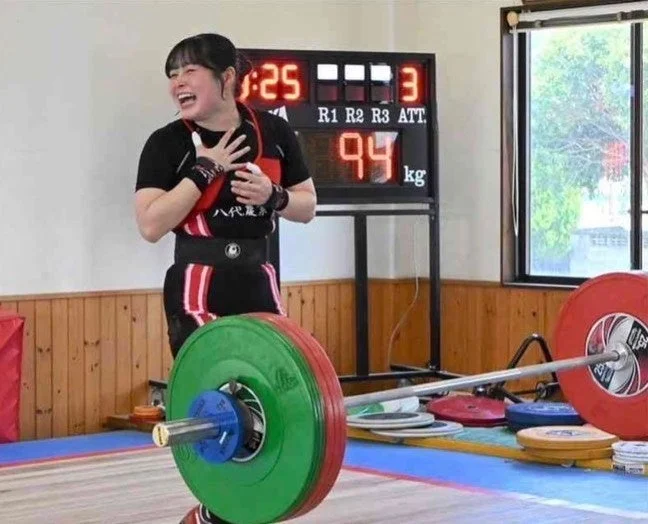 重量挙げの競技中に、94kgのバーベルを挙げた後の女性アスリートが、達成感と喜びの表情を見せている瞬間を捉えた写真です。