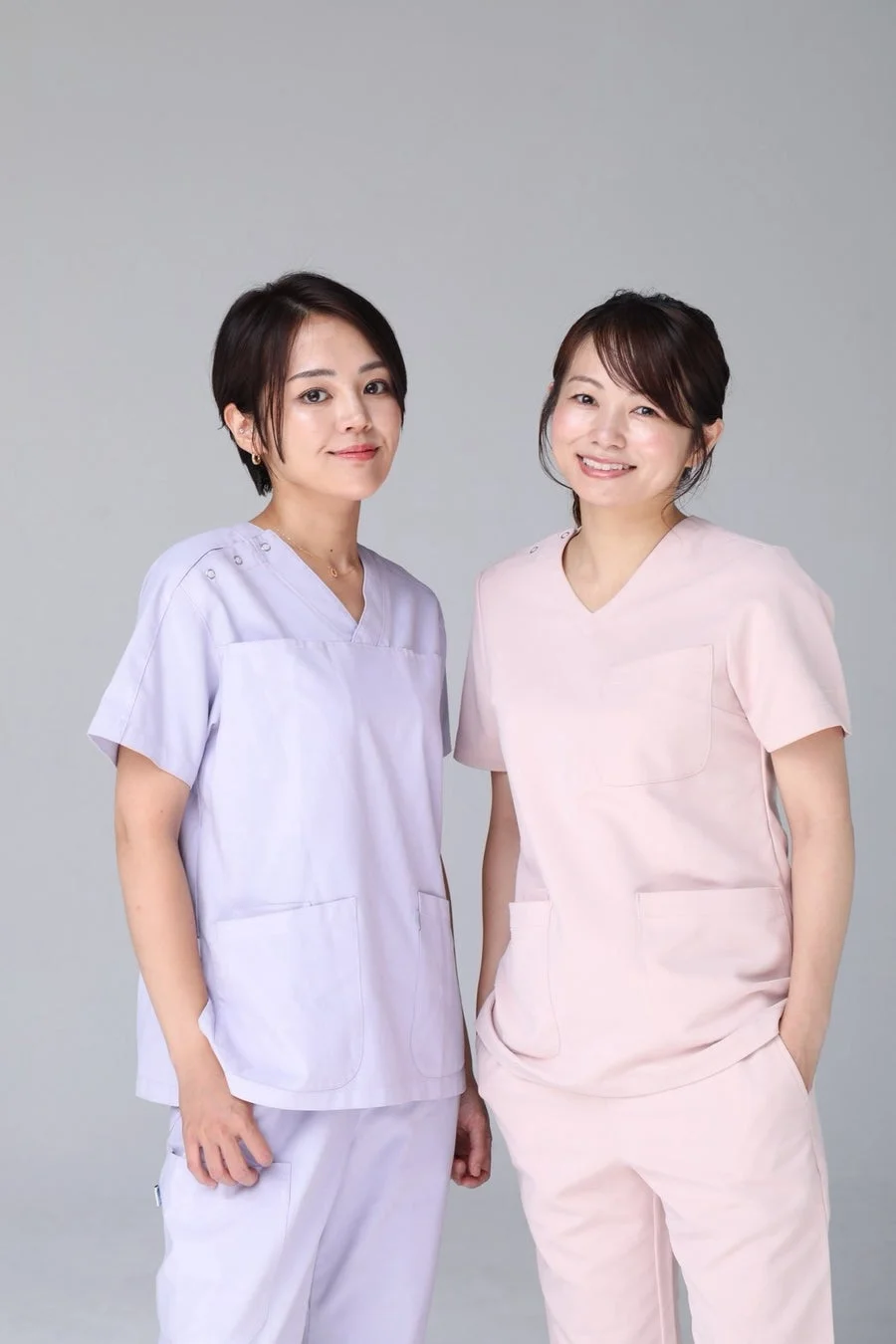 ラベンダーとピンクのスクラブを着た2人の女性が、笑顔で正面を向いているスタジオポートレートです。医療従事者や看護師のイメージを伝えています。