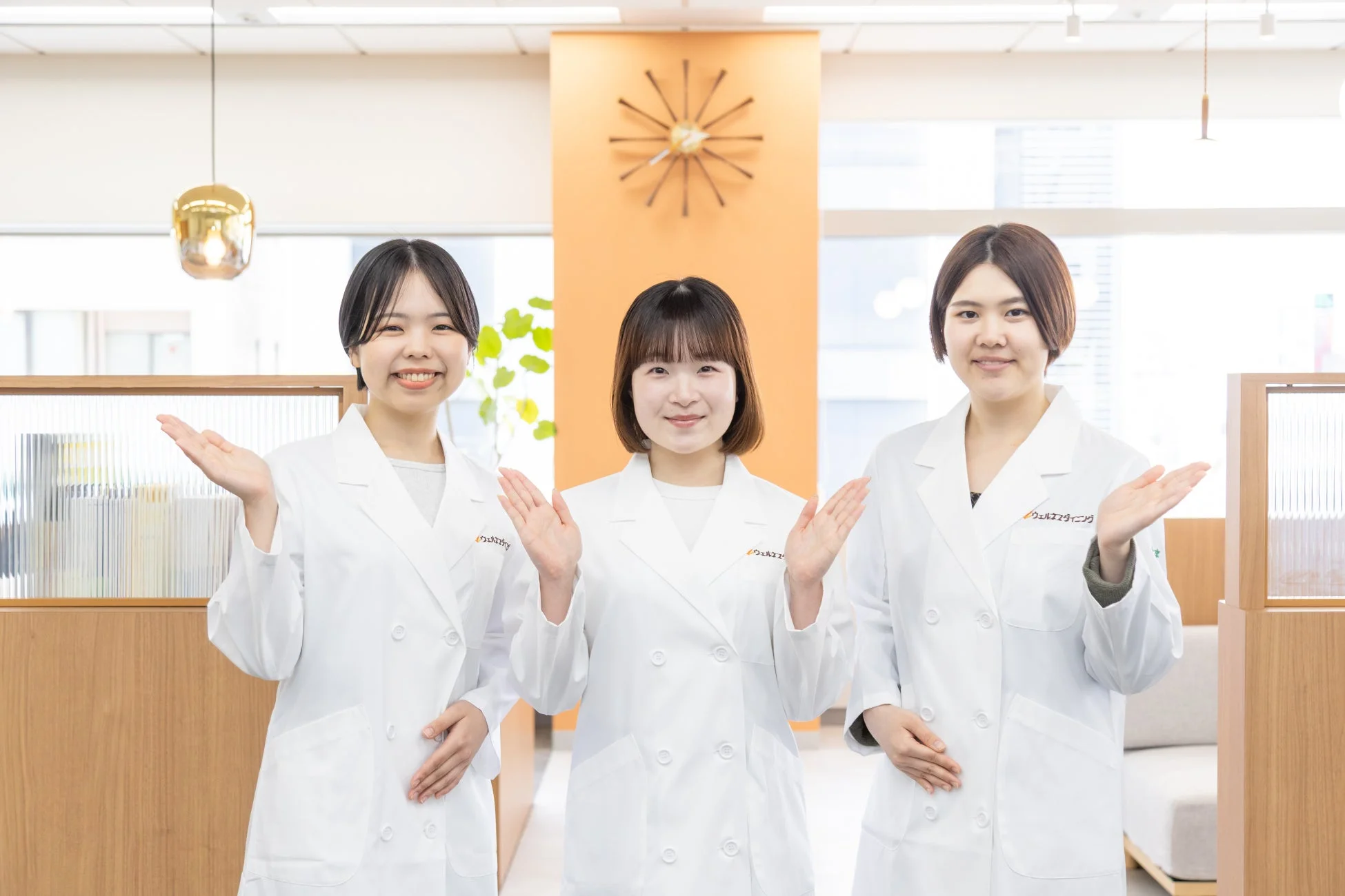 白衣を着た3人の女性が笑顔で両手を広げ、何かを案内している様子です。明るく清潔感のあるオフィスのような空間で、親しみやすいプロフェッショナルな雰囲気が伝わります。