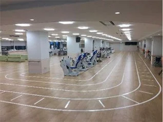 広々とした屋内のフィットネス施設で、木目調の床に白い線でランニングトラックが描かれています。トラックの周りには複数のエクササイズマシンが設置されており、運動やトレーニングに適した空間であることが伺えます。