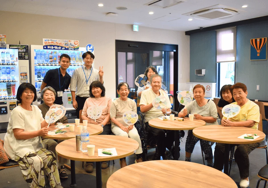 施設内で高齢者と若い男性が交流している様子。参加者は笑顔で団扇を持ち、丸いテーブルを囲んで座っています。和やかな雰囲気のイベント風景です。