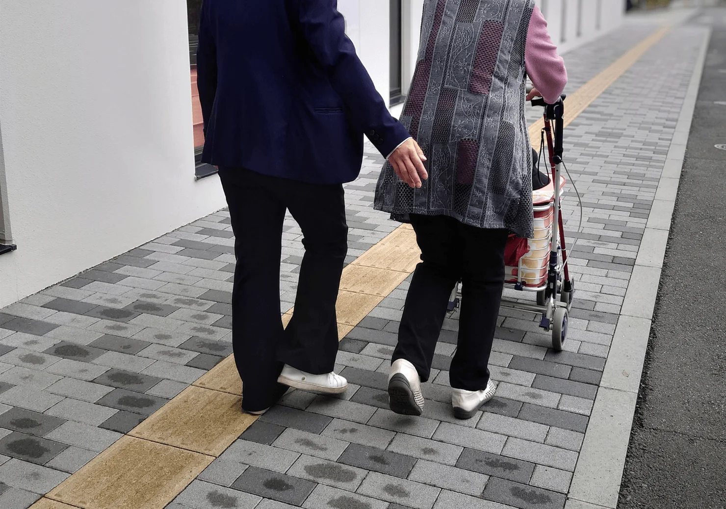 歩行器を使う高齢者と、その隣を歩くもう一人の高齢者が、舗装された歩道をゆっくりと進んでいます。