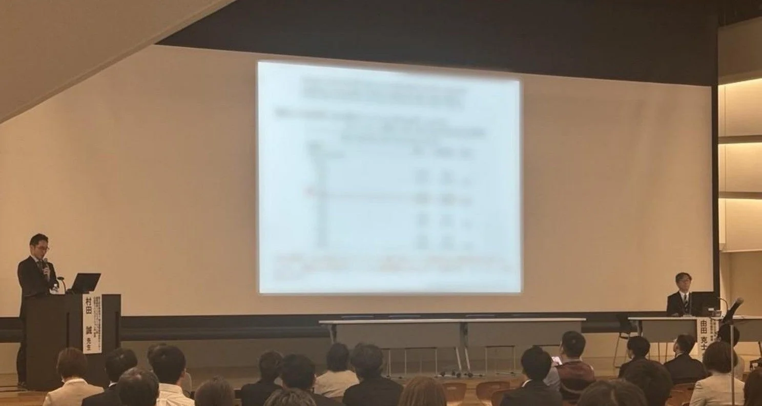 ホールで行われているプレゼンテーションの様子。演台に立つ講演者と、テーブルに着席するもう一人の登壇者、そして大画面に映し出された資料を見つめる多くの聴衆が写っています。