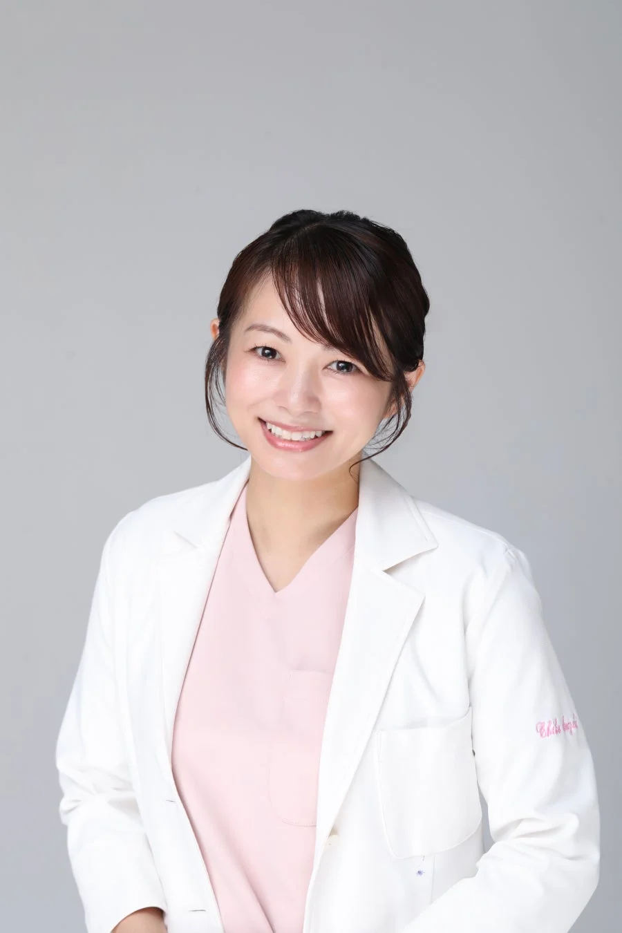 白衣とピンクのスクラブを着用した日本人女性が笑顔でこちらを見ているポートレート。医療従事者か医師のイメージ。