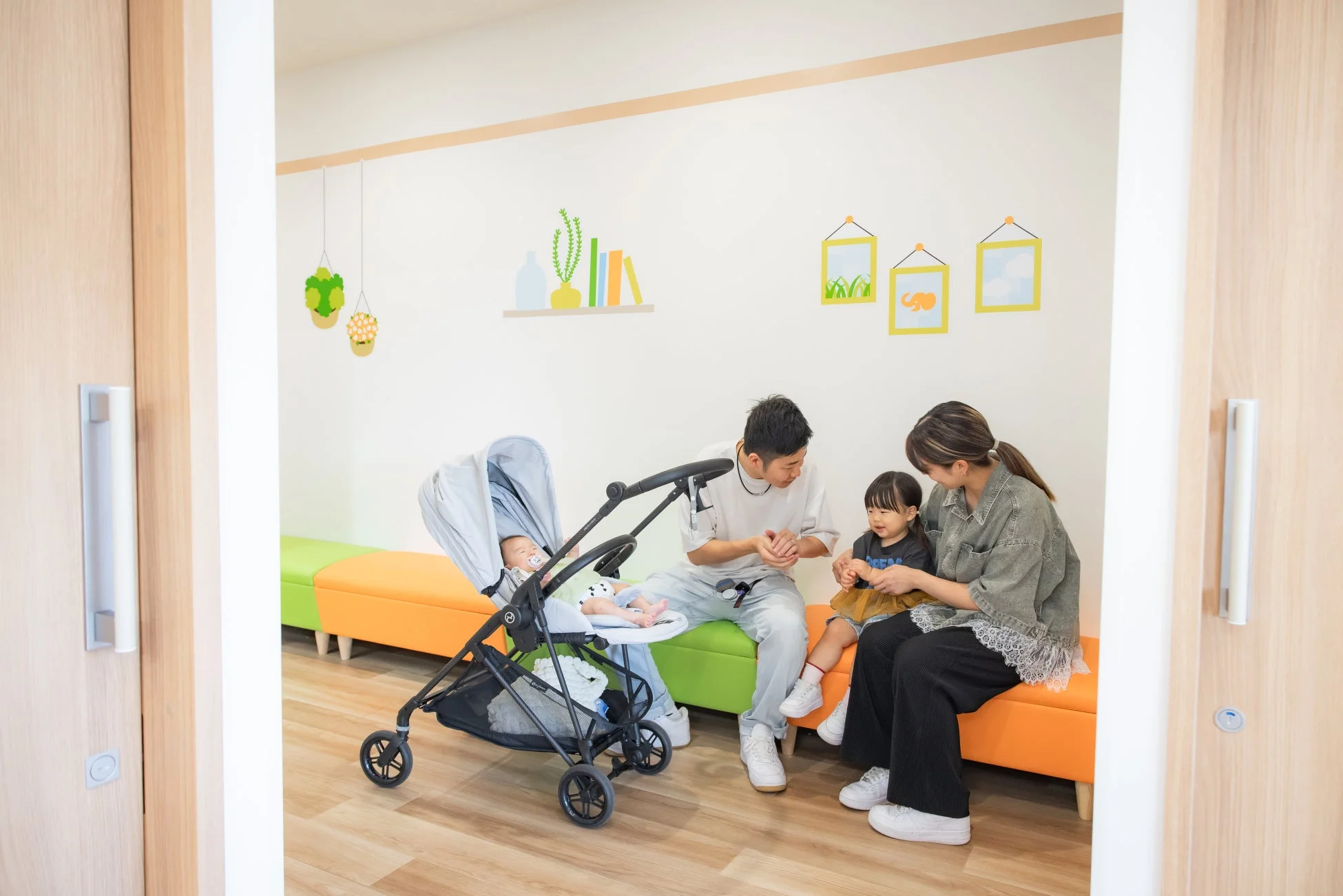 明るい室内で、父親、母親、幼児、ベビーカーに乗った赤ちゃんの4人家族がリラックスしている様子
