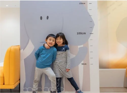 ゾウのイラストが描かれた身長計の前で、男の子と女の子が笑顔で立っている様子