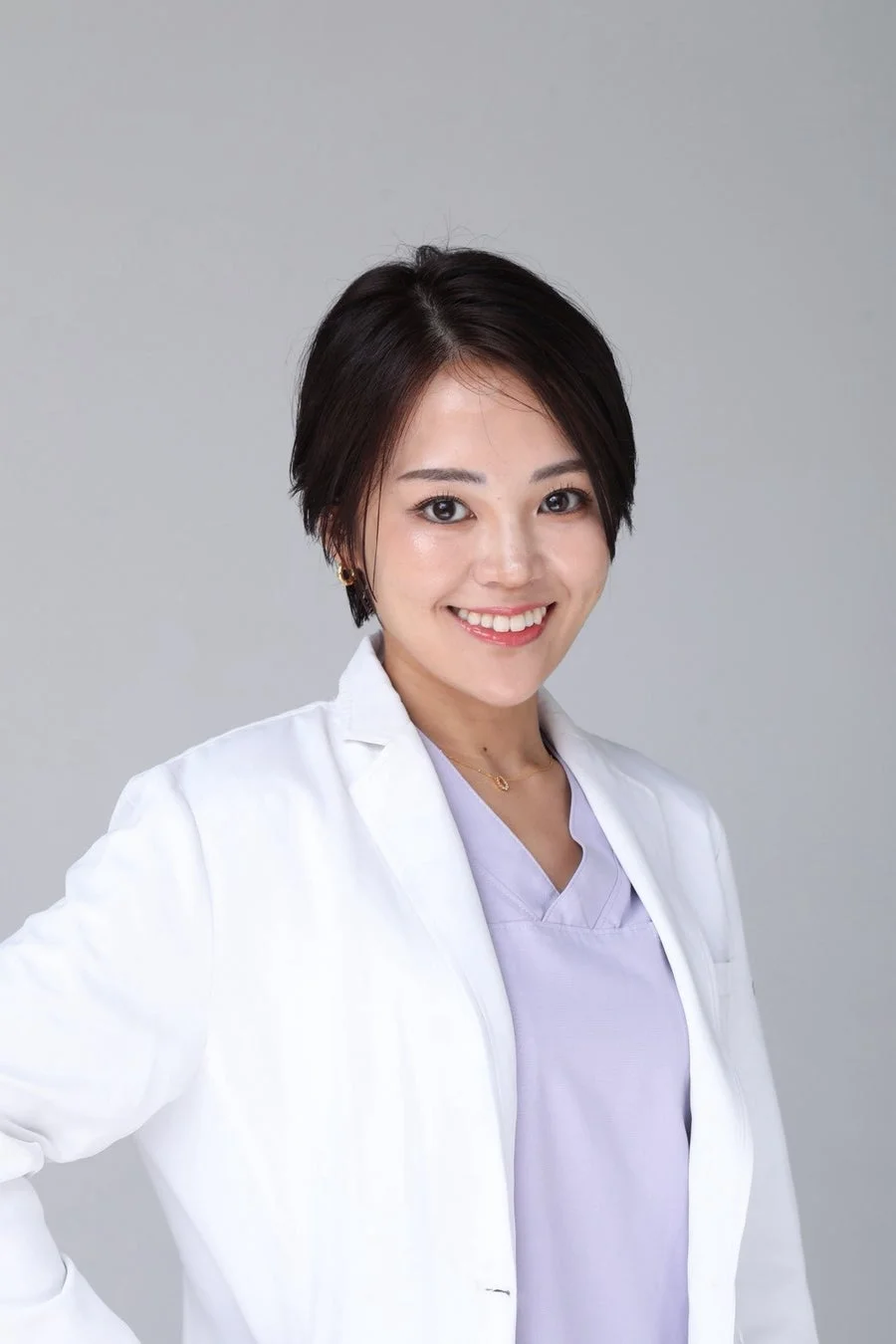 白衣と紫色のスクラブを着用した短髪の女性が、カメラに向かって明るい笑顔を見せているポートレート写真です。清潔感のあるシンプルな背景で、プロフェッショナルな雰囲気が伝わります。