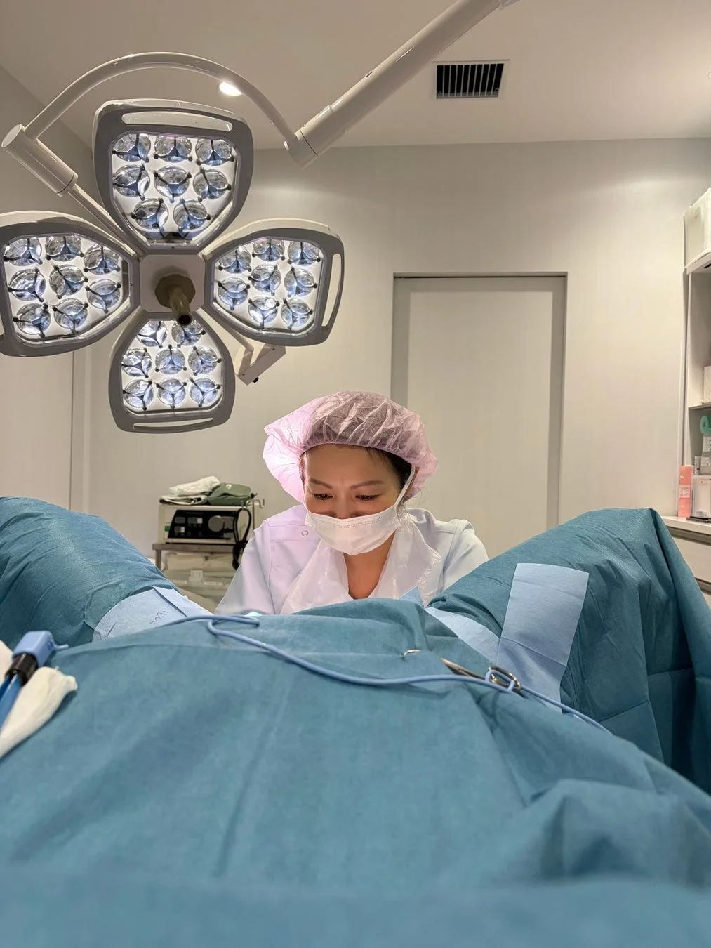 手術室らしき空間で、医療従事者が患者に処置を施している様子。ピンクのキャップとマスクを着用し、患者は青い布で覆われている。上部には大きな手術用ライトが見える。