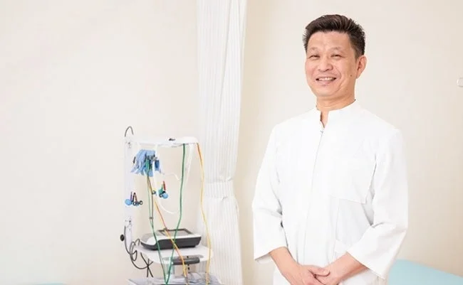 白衣を着たアジア人男性医師が、医療機器が置かれた清潔な診察室で笑顔で立っています。