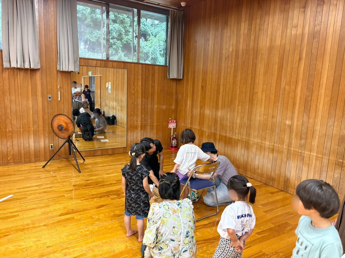 木目調の壁と床の広い室内で、数人の子供と大人が集まって活動している様子