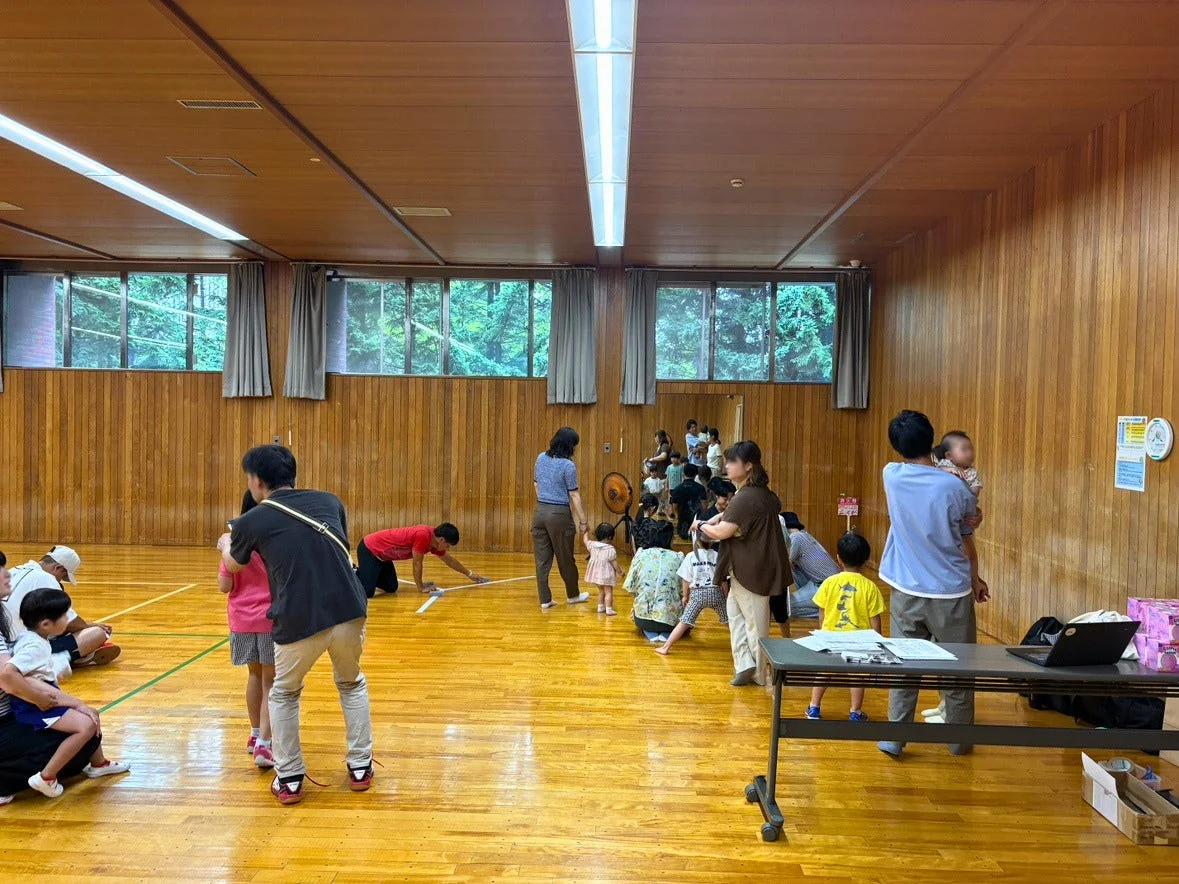 体育館のような広間で、大人と子供たちが集まり、何らかの活動やイベントに参加している様子