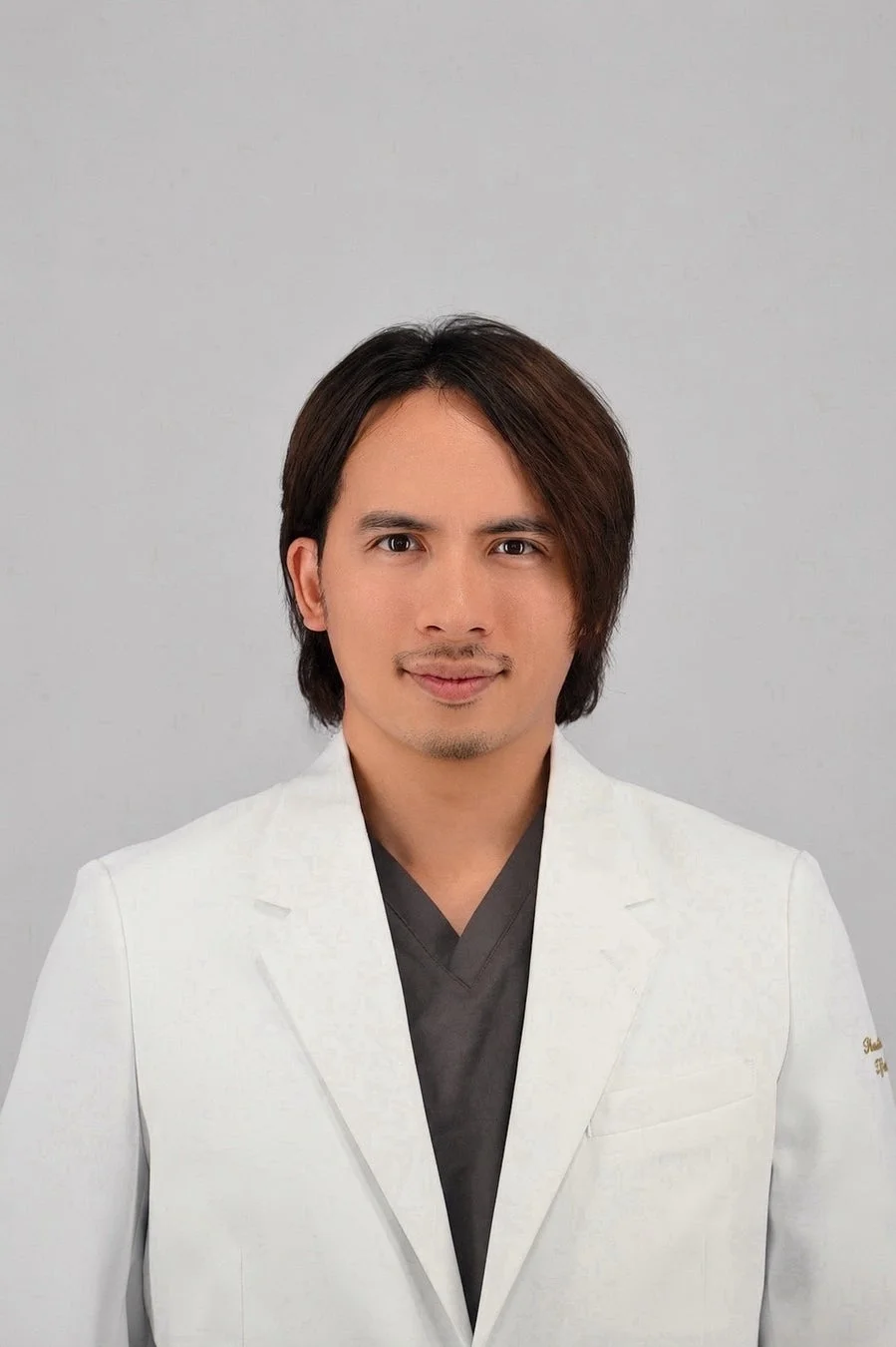 白衣とスクラブを着用した男性のポートレートです。長めの髪と口ひげが特徴で、カメラに向かって穏やかに微笑んでいます。医療関係者か専門家のような印象です。