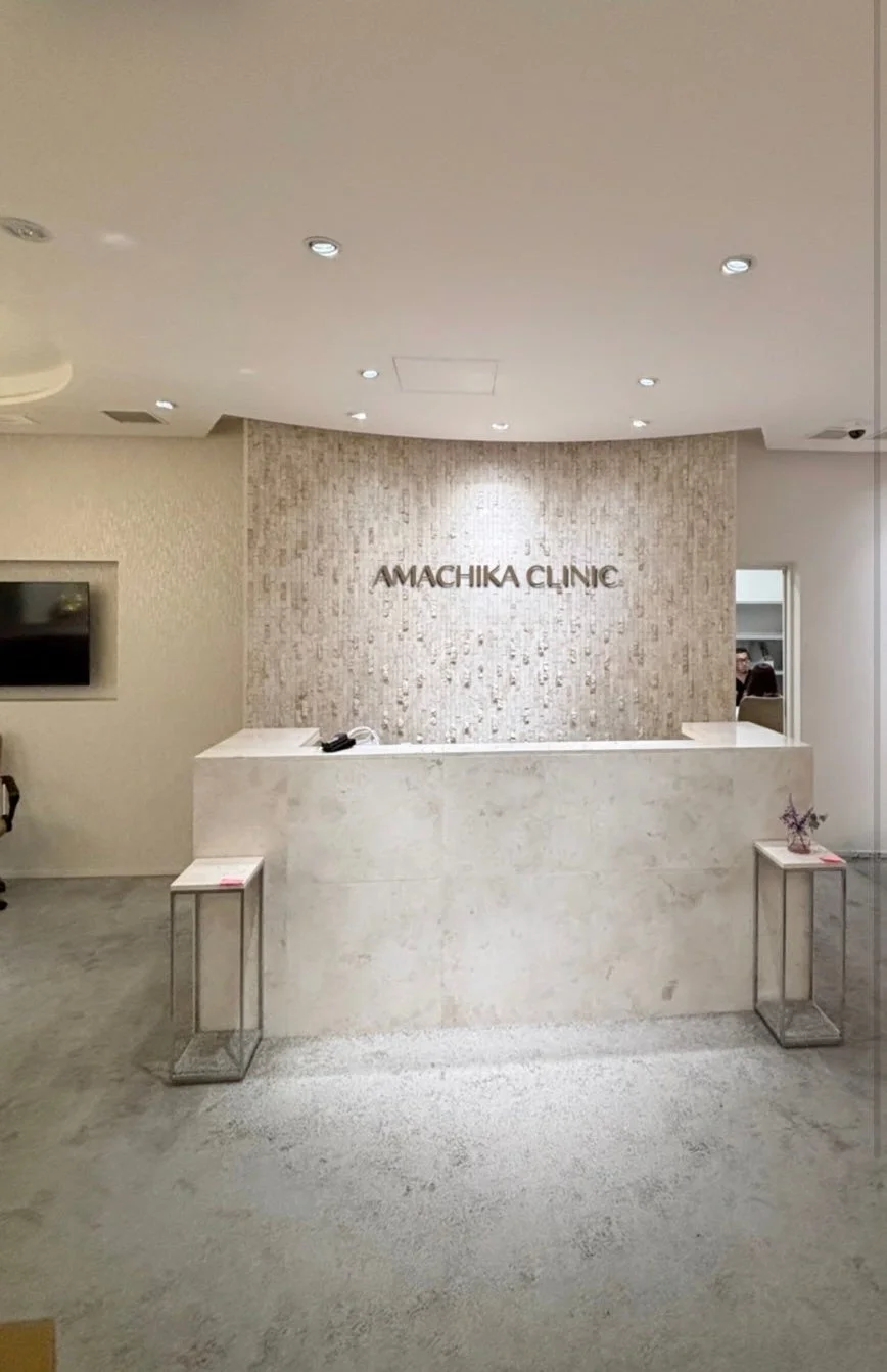 「AMACHIKA CLINIC」の清潔感あふれるモダンな受付エリア。ベージュの石壁と白いカウンターが特徴で、落ち着いた雰囲気が漂う医療機関の内装風景です。