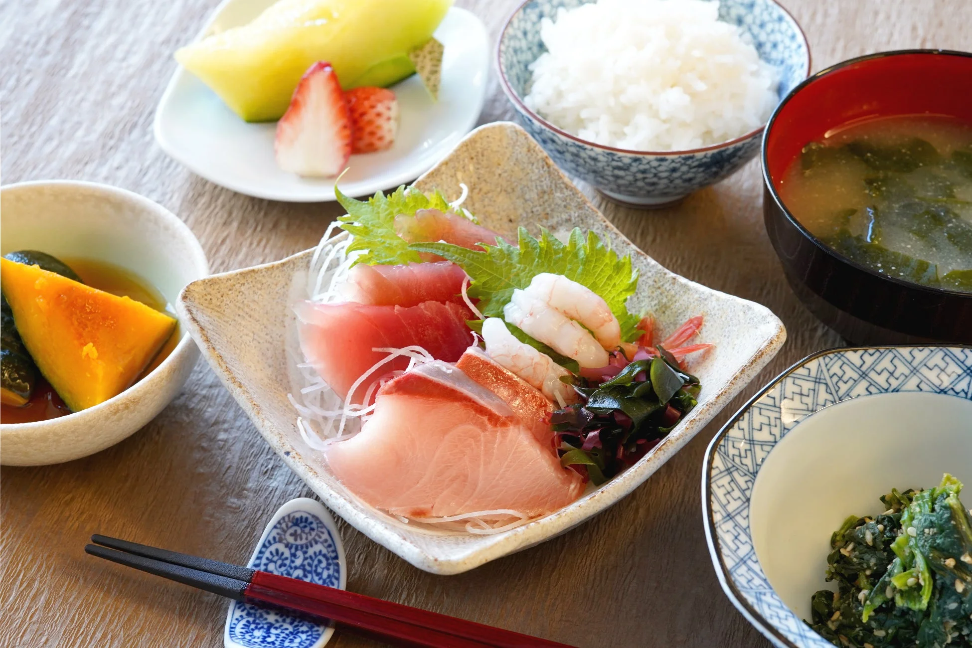 新鮮な刺身盛り合わせを中心に、ご飯、味噌汁、煮物、和え物、デザートのフルーツが並んだ彩り豊かな和定食です。バランスの取れた健康的な食事が食欲をそそります。
