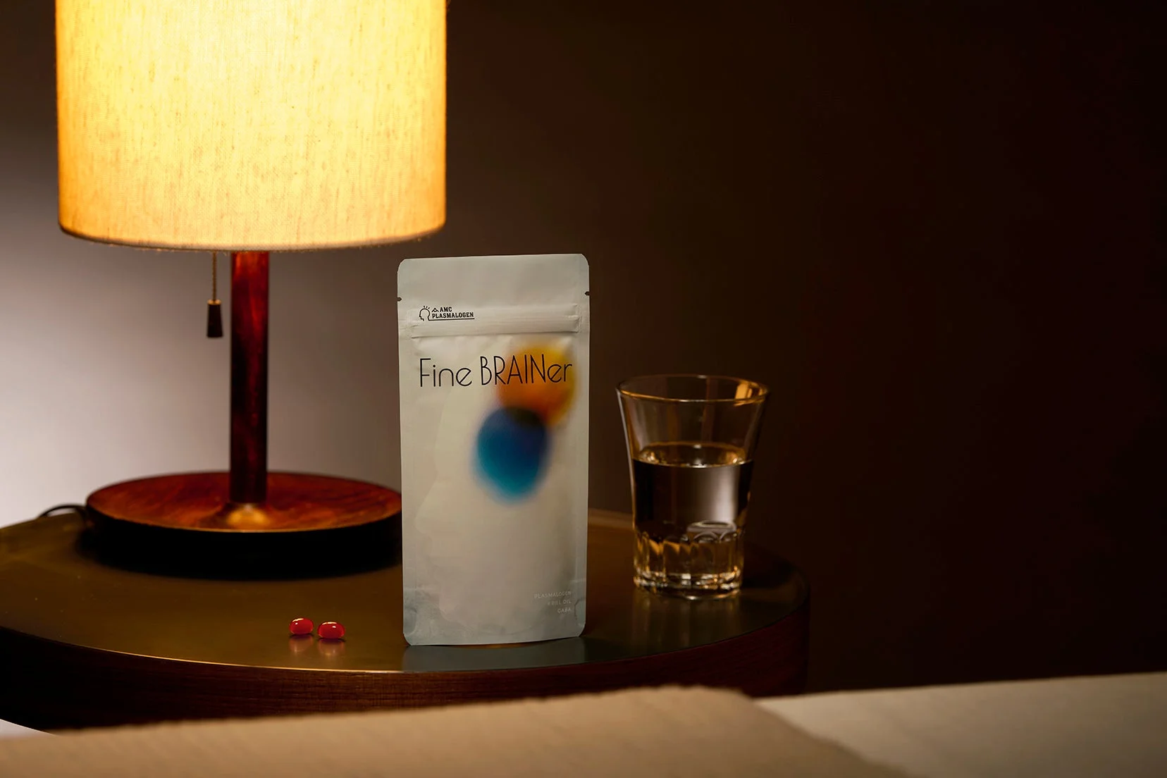 卓上ランプの柔らかな光が灯る部屋で、サプリメント「Fine BRAINer」のパウチ、水、赤い錠剤がテーブルに置かれています。