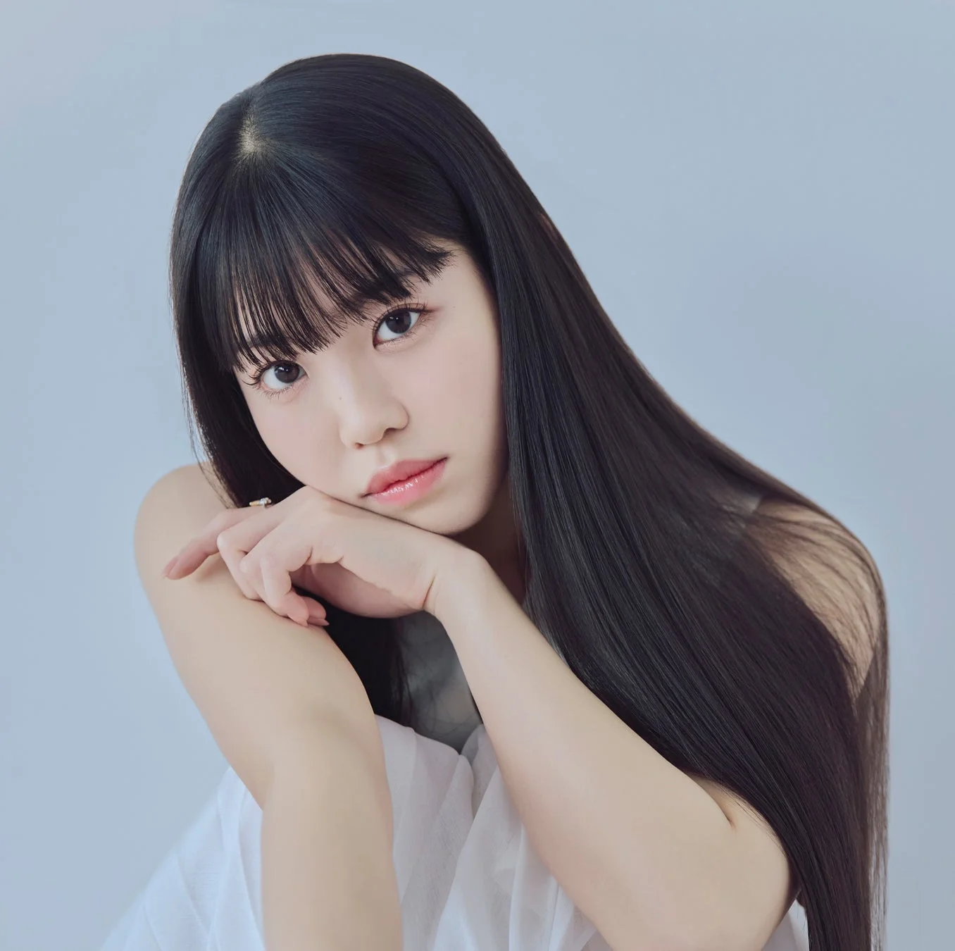 白い衣装を身につけた黒髪ロングヘアの若い女性が、顎に手を添えてカメラを見つめているポートレート写真